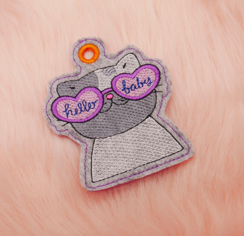 Preview: Stickdatei Set Beach Time Cat Doodle Applikation inkl. ITH Anhänger, Foto zeigt stoffgeösten Anhänger. Katzenbüste in Grautönen, pink-rosa Herz-Brille mit Aufschriften 