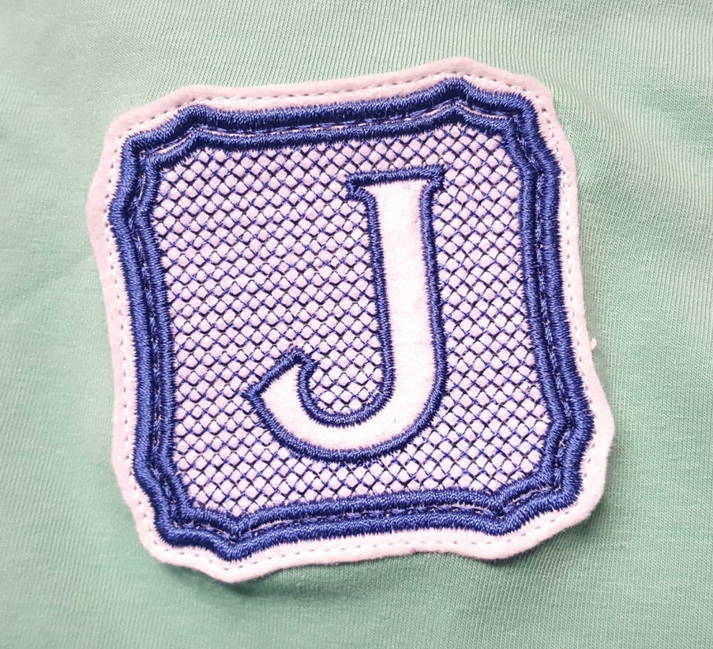 Stickdatei Set Monogramme Elegant geeignet für Frottierwaren, Foto mit Buchstabe "J" in Umrahmung, der auf schilffarbenem Jerseystoff aufgenäht ist. Monogrammfarben sind lila und rosa.
