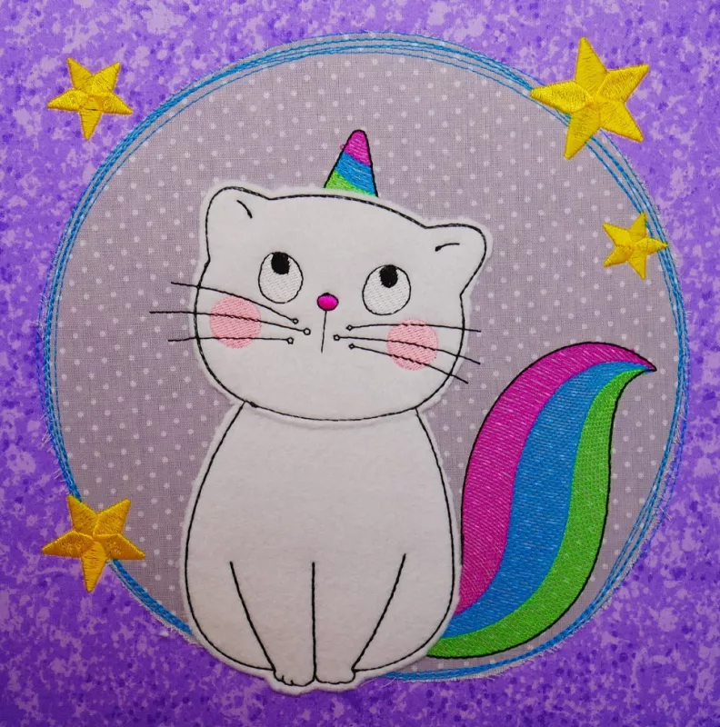 Stickdatei Spar-Set Cat Doodle Applikationen inkl. ITH Anhänger, Foto zeigt Applikation auf lilagemustertem Stoff. Stoffkreis, in dem helle Einhorn-Katze sitzt, Horn und Schwanz bunt. Kreis von gelben Sternen umgeben.