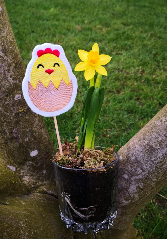 Stickdatei Set Oster-Stabdesigns, Foto mit Ausfertigung in Blumentopf mit Narzisse steckend, vor Baumstammausschnitt im Freien. Küken in halber Eierschale, Farben gelb-rot-schwarz-apricot.