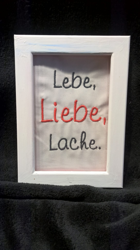 Stickdatei Set Weil ich Dich liebe! (Für 10x10, 13x18 und 18x30), Foto zeigt weißen Bilderrahmen in Hochkantformat. Darin auf hellem Stoff in diversen Farben aufgestickt Schriftzug: "Lebe, Liebe, Lache.".