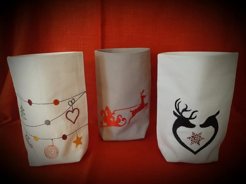 Stickdatei Set 6 winterliche Lichtbeutel (für den 13x18 Rahmen), Foto mit 3 Beuteln aus hellem Stoff. 1 mal Santa-Rentier-Schlitten, 1 mal herzförmig einander zugewandte Rentiere, 1 mal Schnüre mit Weihnachtssymbol-Behang.