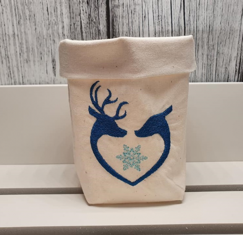 Stickdatei Set 6 winterliche Lichtbeutel (für den 13x18 Rahmen), Foto mit Beutel aus hellem Stoff. Bestickung mit 2 dunkelblauen einander zugewandten Rentierköpfen in Herzform. Zwischen ihnen heller Eiskristall.