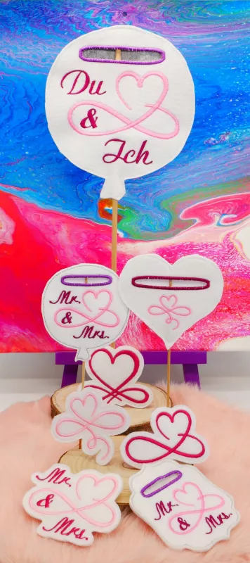 Stickdatei Set Liebe & Hochzeit - Stabdesigns, Foto mit 8 Designs, auf einer Holzscheibe präsentiert, Untergrund oranges Fell, bunter Hintergrund. Mit Einstecköse: 2 mal Luftballons ("Mr. & Mrs."; "Du & Ich"); 1 mal Herz mit Herzstick; 1 mal Schnörkelrahm