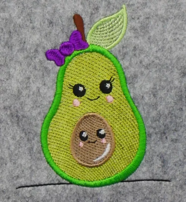 Stickdatei Set Sweet Avocados, Foto mit einem Stickdesign auf graumeliertem Filz. Die grüne Avocado hat ein pausbäckiges Gesicht, ein grünes Blatt und eine lila Schleife, im Inneren ist ein brauner Kern mit Pausbacken-Gesicht.