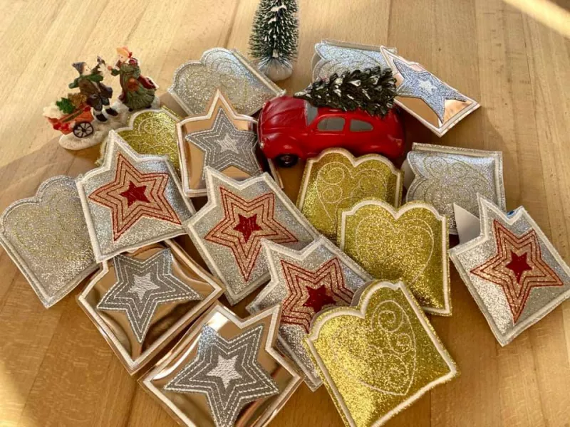 Stickdatei Set ITH Gutschein- und Schokohüllen Danke und Geburtstag. Foto zeigt auf Holzboden viele quadratische Ausfertigungen, je Stern- oder Herzmotiv, dazwischen als Dekozusatz Nippes in Weihnachtsoptik. Oberer Hüllenrand ist der Form des Motivrandes 