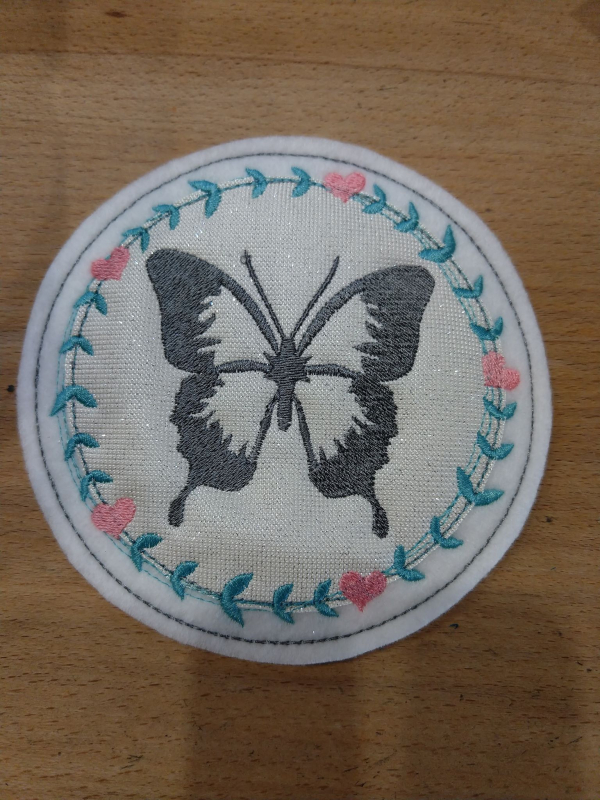 Stickdatei Set Hallo Frühling (div. Rahmengrößen), Foto zeigt kreisrunden Mug Rug. Auf hellem Stoff mittig dunkelgrau-weißer Schmetterling, umgeben von Kranz aus Herzen und Blättern.