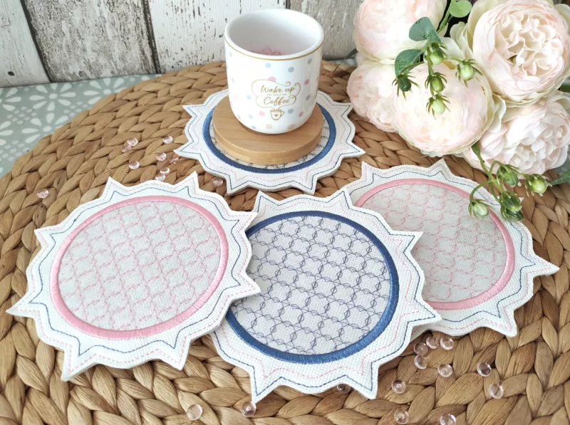 Stickdatei Set ITH Mug Rug 6, Ausschnitt eines Tischarrangements mit Platzdeckchen, auf dem vier Mug Rugs liegen. Auf einem davon befindet sich ein Holzuntersetzer, auf dem eine bedruckte Tasse steht. Daneben ein helles Blumenarrangement. Alle Mug Rugs ha