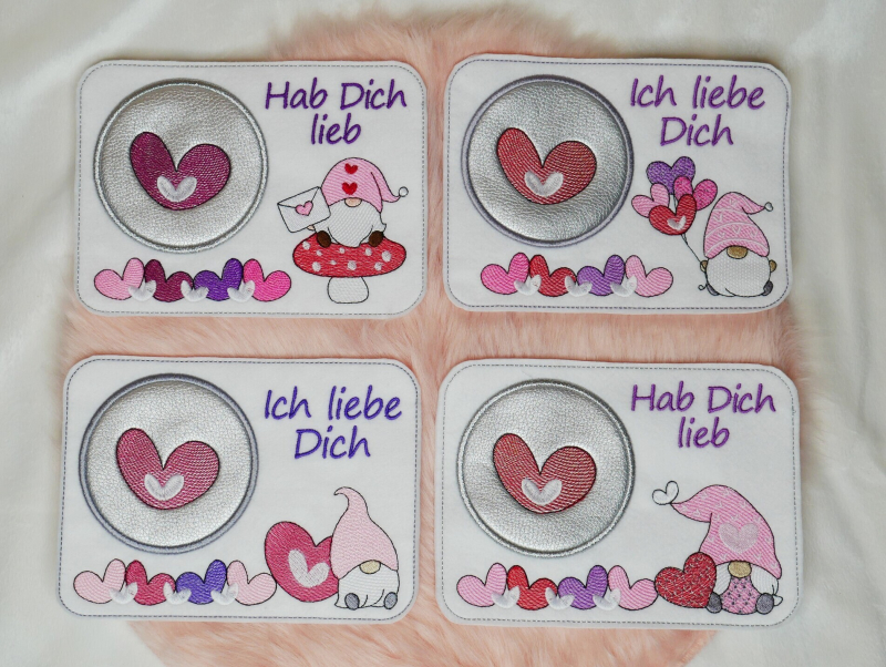 Preview: Stickdatei Set Lovegnomes ITH Mugrugs, Foto mit 4 Ausfertigungen, auf apricotfarbenem Fell liegend.hellem Untergrund mit Becher, Holzherz und Blattzweig als Dekobeigabe. Rechteckige helle Tassenteppiche mit unterschiedlichen bunten Gnommotiven.