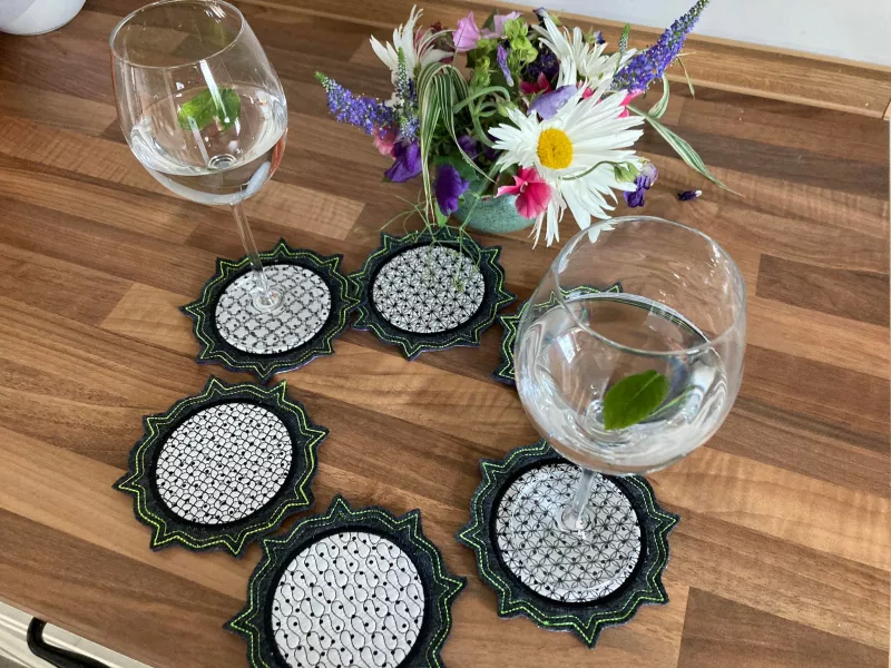 Stickdatei Set ITH Mug Rug 6, Tischarrangement mit kleinem buntem Blumenstrauß und sechs kreisförmig ausgelegten Mug Rugs, wobei zwei von ihnen je ein Weinglas tragen. Alle sechs haben einen strahlenförmig und doppelt am Rand abgesteppten dunkelgrauen Fi