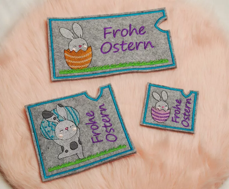 Stickdatei Set Osterhüllen ITH Geschenk- und Schokoladenhüllen + Einzelmotive, Foto mit 3 Ausfertigungen, die auf orangem Fell liegen. Unterschiedliche rechteckige Formate aus grauem Filz, bestickt mit diversen Hasenmotiven und Schriftzug "Frohe Ostern".