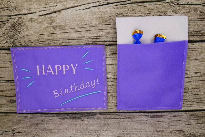 Stickdatei Set ITH Geburtstagskarten (10x10 u. 13x18), Foto mit 2 lila Karten. Karte im Hochkantformat zeigt ihre Rückseite, die eine Einstecktasche aufweist, aus der 2 Bonbonpapierspitzen ragen.Karte im Querformat zeigt Bestickung: "HAPPY Birthday!"/Str