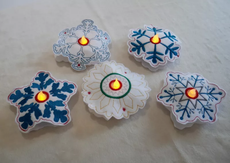 Stickdatei Set ITH Schneeflocken in 2 Größen, Foto zeigt 5 Schneeflocken, auf hellem Untergrund präsentiert. Jede im Zentrum LED-Licht-beleuchtet. Stickornamente divers, hauptsächlich blau-weiß.