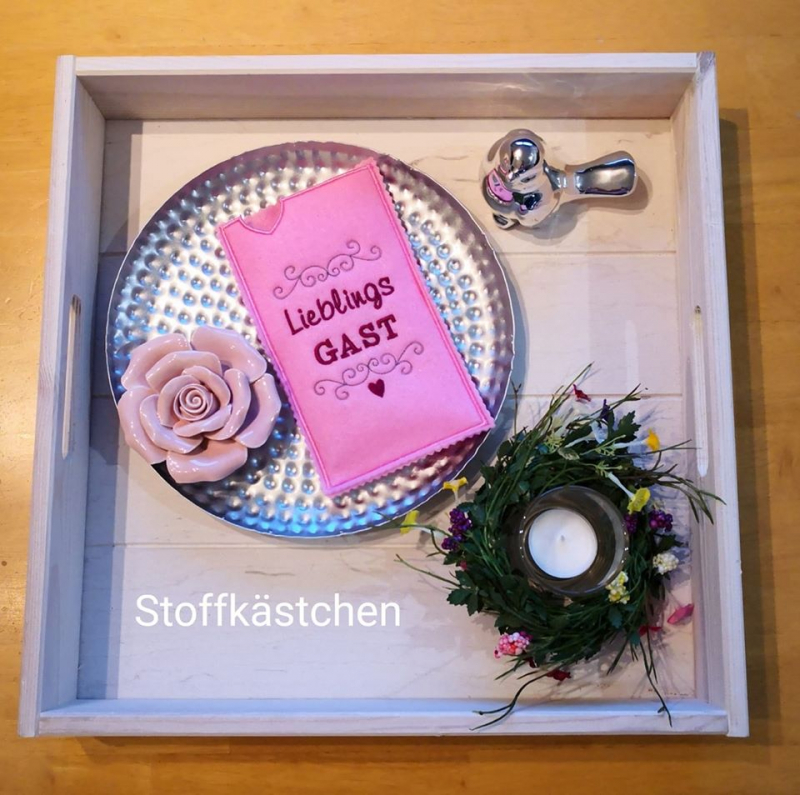 Stickdatei Set ITH Geschenkhüllen für Schokolade, Gutscheine u.a., Foto mit Hochkant-Hülle in Pink, die auf einem Tablett mit Deko und Teller hübsch platziert ist. Dunkelpinke Aufschrift "LieblingsGAST" mit Ornamenten und Herz.