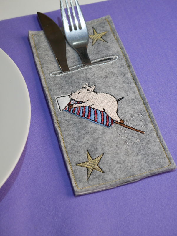 Stickdatei Set Happy new Year Tischdeko (div. Rahmengrößen) , Foto mit länglicher Bestecktasche, gefüllt mit Messer/Gabel, auf lila Untergrund. Tasche aus grauem Filz, Bestickung: Schwein auf Rakete, Sterne.