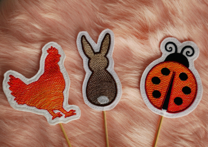 Stickdatei Set ITH Frühlings-Stabdesigns, Foto mit 3 Designstäben, auf orangem Fell liegend. Motive: Huhn (rot), Hase (braun-weiß), Marienkäfer (rot-schwarz).