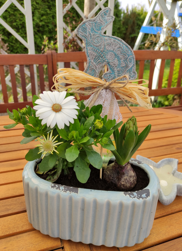 Stickdatei Sparset ITH Ostermandala-Deko,
Foto mit in Blumentopf steckendem Dekostab mit Kaninchen, Schleife am Stab. Topf steht auf Tisch, Hintergrund lässt Garten erahnen. Kaninchen aus grauem Filz, welcher filigran türkis bestickt ist.