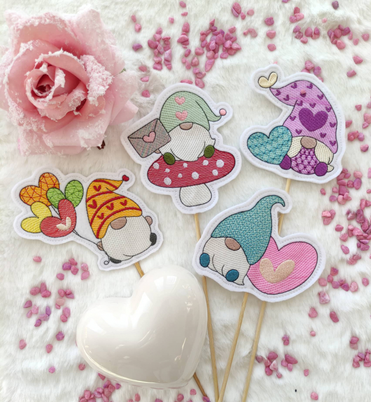 Stickdatei Set Lovegnomes Sparset 4 in 1 mit Dateien für Mugrugs, Schokohüllen, Anhänger und Stabdesigns. Foto mit 4 Stabdesigns, auf hellem Untergrund mit rosa Steinchen liegend, Rosenblüte und Herz als Deko-Beilage. Bunte Gnom-Designs: Gnom mit Herzluft