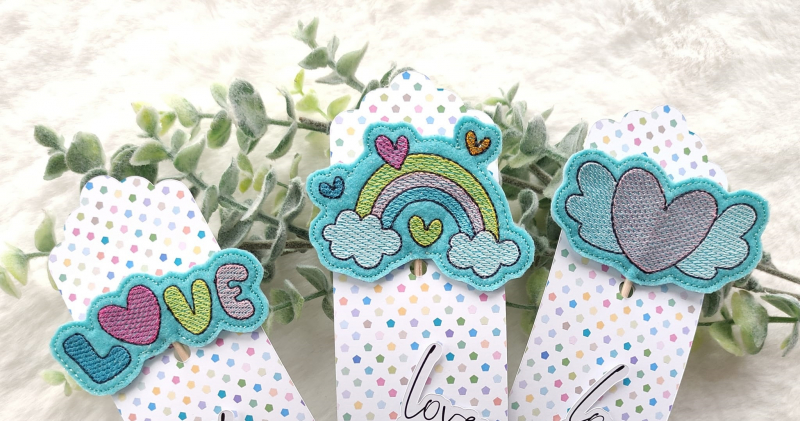 Stickdatei Set Valentinsmöpse ITH Stabdesigns für Stabdeko, Foto mit drei Variationen auf türkisem Filzuntergrund, drapiert auf grünem Zweig: Bunter Schriftzug "LOVE" mit Herz als "O", bunter Regenbogen mit je einer hellen Wolke an den Enden, und umgebend