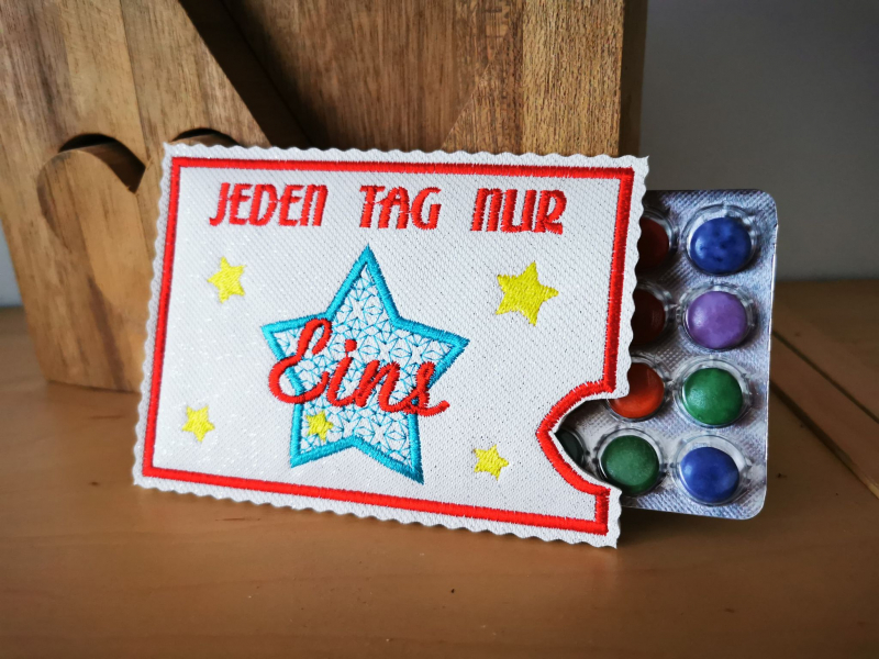 Stickdatei Set ITH Hüllen für Adventskalender to go, Foto mit heller querformatiger Hülle, aus der ein Blister mit bunten Dragees ragt. Bestickung in rot-gelb-hellblau, Sterne, Aufschrift "JEDEN TAG NUR Eins".