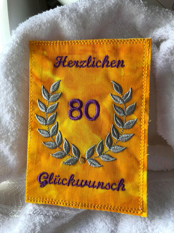 Stickdatei Set ITH Glückwunschkarten inkl. Abwandlung zur Geschenktasche (div. Rahmengr.). Foto zeigt Hochkant-Karte aus orangegelbem Stoff. Oben offener Blätterkranz in der Mitte der Karte, darin die Zahl "80". Schriftzug: "Herzlichen Glückwunsch".