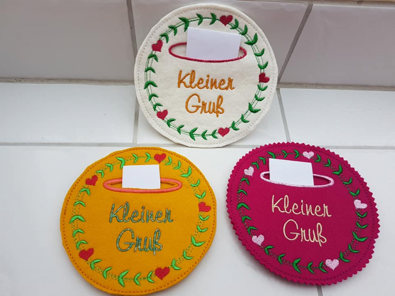 Stickdatei Set Hallo Frühling (div. Rahmengrößen), Foto zeigt 3 kreisrunde Ausfertigungen unterschiedlicher Farben mit Einstecktasche. Kranz aus Herzen und Blättern. Schriftzug "Kleiner Gruß".