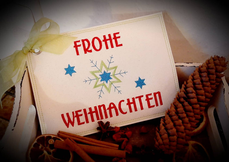 Stickdatei Set ITH Weihnachtskarten, Geschenkanhänger und Schriftzüge, Foto zeigt eine Karte in einer Kiste mit Weihnachtsdekoartikeln. Weiße, metallgeöste Karte mit Bestickung Sterne/Eiskristall, Aufschrift "Frohe Weihnachten".