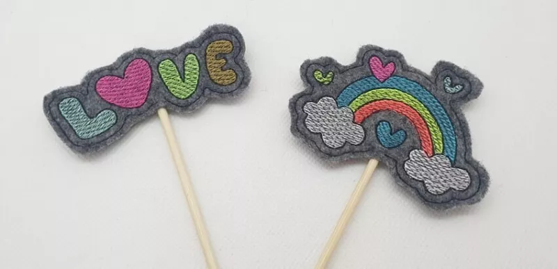 Stickdatei Set Valentinsmöpse ITH Stabdesigns für Stabdeko, Foto mit zwei Designs auf jeweils anthrazitfarbenem Filzuntergrund. Einmal bunter Schriftzug "LOVE" mit rotem Herz als "O", einmal bunter Regenbogen, von Herzen umgeben und mit je einer Wolke