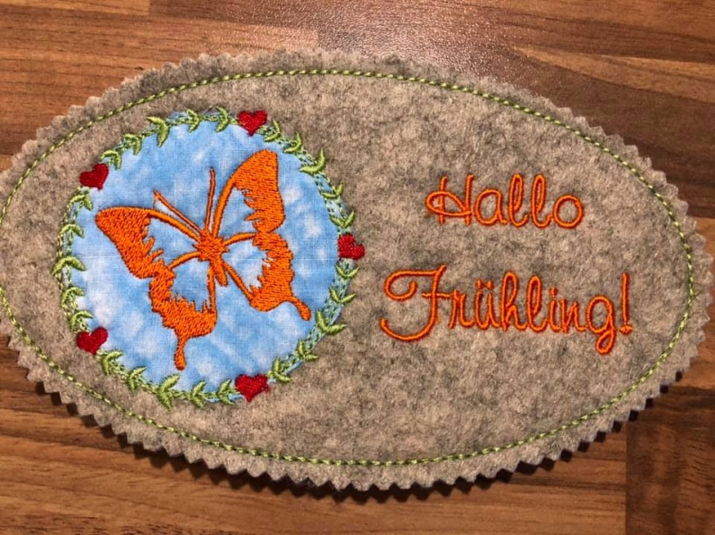 Stickdatei Set Hallo Frühling (div. Rahmengrößen), Foto zeigt ovalen Mug Rug aus Filz in bronzefarben. Links oranger Schmetterling auf rundem hellblauem Grund, umgeben von Kranz aus Herzen und Blättern. Rechts Schriftzug "Hallo Frühling!".