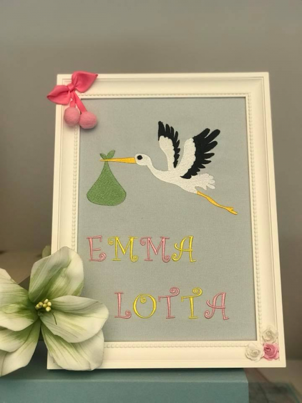 Stickdatei Set Hello Baby (div. Größen), Foto zeigt Bilderrahmen mit eingespanntem gräulichem Stoff. Dieser ist bestickt mit einem Klapperstorch mit Stoffbeutel im Schnabel. Darunter Schriftzug "Emma Lotta".