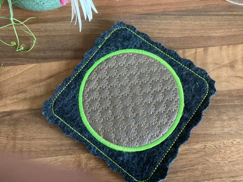 Stickdatei Set ITH Mug Rug 10. Der Ausschnitt zeigt auf einem Holzuntergrund  den Mug Rug als quadratischen dunkelgraublauen wellig geschnittenen Filz mit hellgrün abgestepptem Rand. Mittig ein wellig-blattartiges Ornament in grau, das mit dem hellgrünen