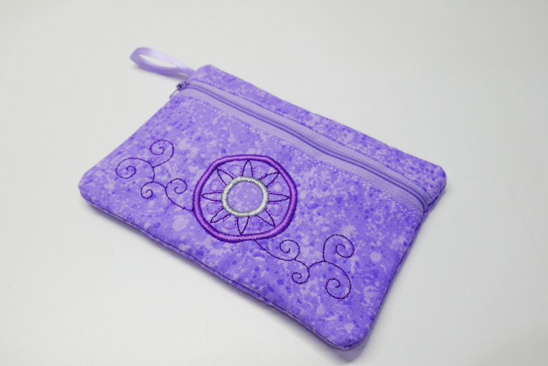 Stickdatei Set BLANKO ITH Taschen hochkant mit Reißverschluss, Foto auf hellem Untergrund mit rechteckiger querformatiger Tasche mit lila Reißverschluss im oberen Teil der Front und lila Stoffschlaufe an der linken Ecke. Stoff lila gemustert mit unten kre