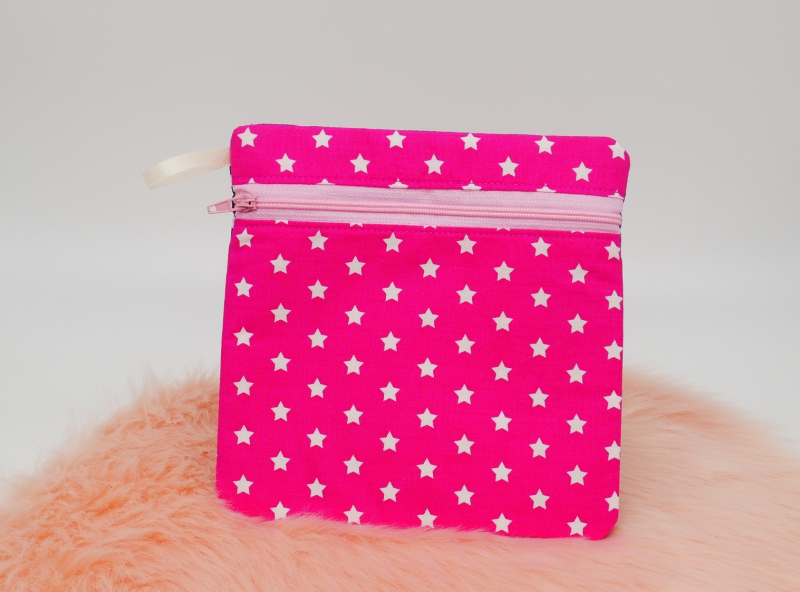 Preview: Stickdatei Set BLANKO ITH Taschen mit Reißverschluss über 60 Größen, Foto zeigt auf orangefarbenem Fell platzierte quadratische Tasche. Rosa Reißverschluss frontal oben, daneben helle Stoffschlaufe an einer Ecke. Bedruckter Stoff in pink mit hellen Sterne