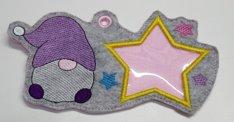 Stickdatei Set ITH Geschenk- und Schokoladenhüllen mit Fenster, Foto zeigt Ausfertigung eines länglichen, mittig geösten Anhängers, der auch als Hülle genutzt werden kann. Gestaltung: auf grauem Filz ein Gnom, daneben großer Stern aus Klarsichtmaterial