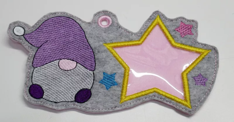 Stickdatei Set ITH Geschenk- und Schokoladenhüllen mit Fenster, Foto zeigt Ausfertigung eines länglichen, mittig geösten Anhängers, der auch als Hülle genutzt werden kann. Gestaltung: auf grauem Filz ein Gnom, daneben großer Stern aus Klarsichtmaterial