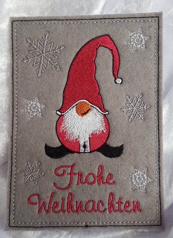 Stickdatei Set ITH Glückwunschkarten inkl. Abwandlung zur Geschenktasche (div. Rahmengr.), Foto zeigt Hochkant-Karte aus mittelgrauem Filz. Gnommotiv, hauptsächlich rot, umgeben von hellen Sternen und rotem Schriftzug "Frohe Weihnachten".