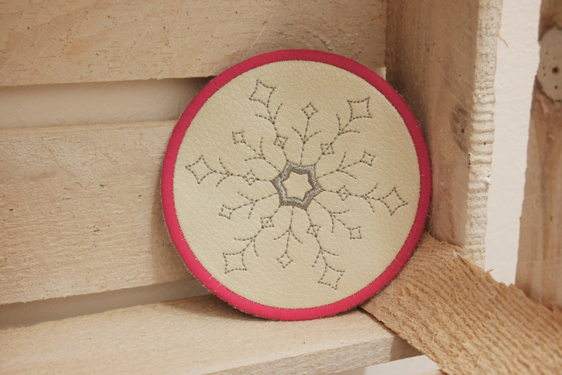 Freebie Stickdatei ITH Mug Rug Winterzeit (13x18 und 10x10 Rahmen), kreisrund aus beigem Kunstleder, rot umrandet. Motiv innen grauer filigraner Eiskristall.