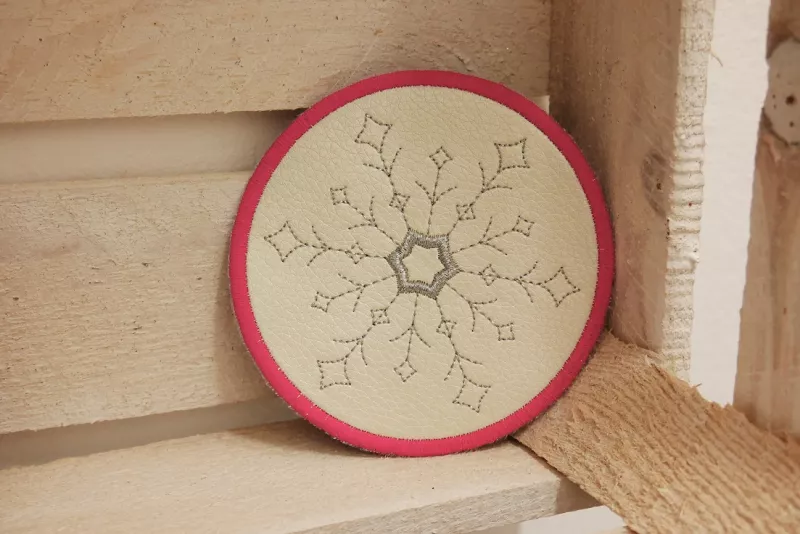 Freebie Stickdatei ITH Mug Rug Winterzeit (13x18 und 10x10 Rahmen), kreisrund aus beigem Kunstleder, rot umrandet. Motiv innen grauer filigraner Eiskristall.