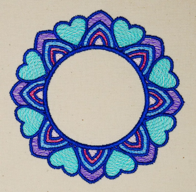 Stickdatei Set Mandalas + Split Mandala (div. Größen), Foto von Ornament auf hellem Stoff. Buntes Mandala-Ornament in blau-rot-lila aus Herzen und abgerundeten Zacken mit kreisrundem, freiem Zentrum.