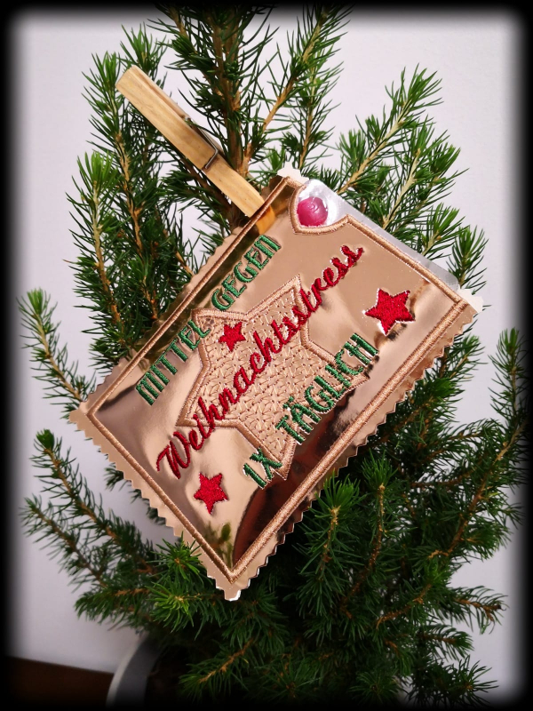 Stickdatei Set ITH Hüllen für Adventskalender to go, Foto zeigt querformatige Hülle, mit Klammer an Nadelbaum befestigt. Glänzende Grundlage, rot und grün gestickter Schriftzug "Mittel gegen Weihnachtsstress- 1 x TÄGLICH".