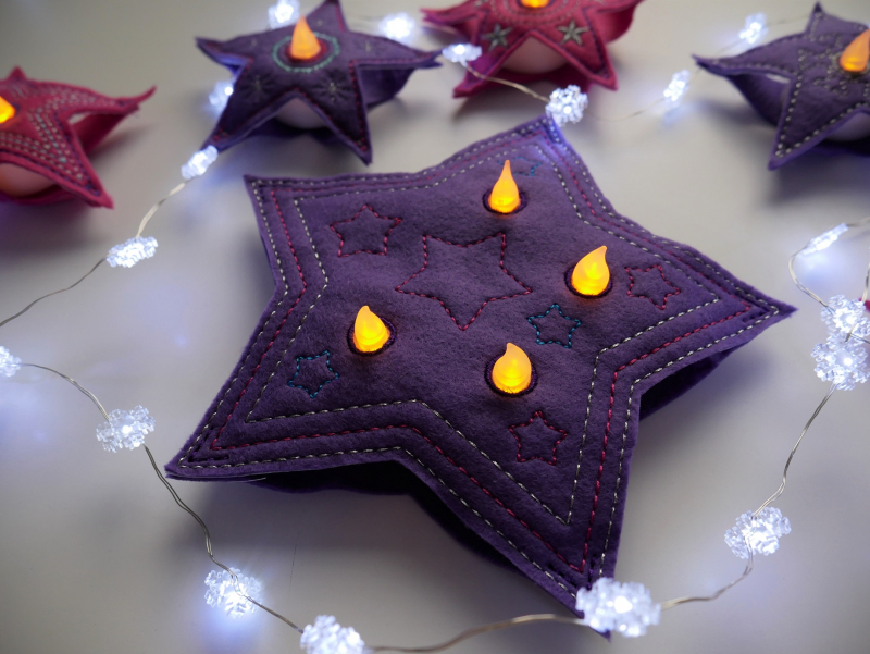 Stickdatei Set Christmas Lights (div. Rahmengrößen), Foto zeigt als LED-Cover in Sternformat 1 großen lila Stern mit 4 Öffnungen für Licht, in die leuchtende LEDs eingesteckt sind. Umgeben im Hintergrund von kleineren Sternen ähnlicher Machart.