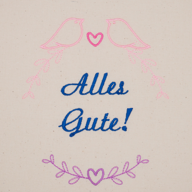 Stickdatei Set florale Rahmen + Schriftzüge, Foto mit Motiv in lila, pink, rosa, dunkelblau auf hellem Stoff. Begrenzung durch 3 Blattranken mit Herzen, oben zwei Vögel, mittig Schriftzug "Alles Gute!".