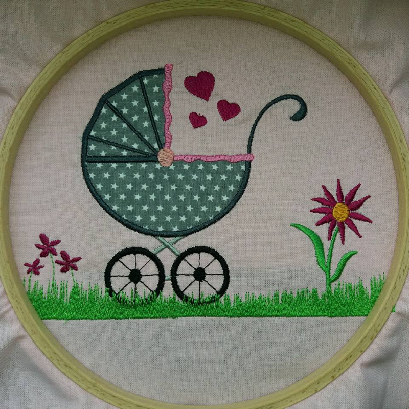 Stickdatei Set Kinderwagen Applikation u. Vollstick, Foto zeigt runden Stickrahmen mit hellem eingespanntem Stoff. Innerhalb des Rahmens in bunt Kinderwagen mit Stoffapplikation auf Wiese, Herzchen, Blumen. 