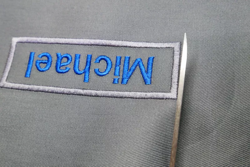 Stickdatei Set Blanko-Patches Aufnäher (mit und ohne Klett auf der Rückseite umsetzbar). Foto zeigt, wie ein fertiger länglicher Aufnäher auf grauem Stoff, blauer Schriftzug "Michael", mit einer Schere ausgeschnitten wird.