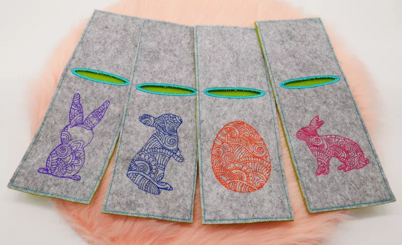 Stickdatei Sparset ITH Ostermandala-Deko, Foto mit 4 nebeneinander auf orangem Felluntergrund liegenden Hochkant-Bestecktaschen aus grauem Filz mit grün umrandeter Einstecköffnung. Motive: 3 mal unterschiedliche Kaninchen/Hasen; 1 mal Osterei. Alles filig