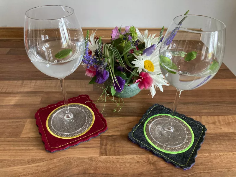 Stickdatei Set ITH Mug Rug 10. Zum Einen dunkelroter, wellig geschnittener quadratischer und am Rand abgesteppter Filz mit rundem grauem Stickemblem, welches kreisförmig mit gelbem  Garn umstickt ist. Zum Anderen Filz in dunkelgraublau mit ebenfalls graue
