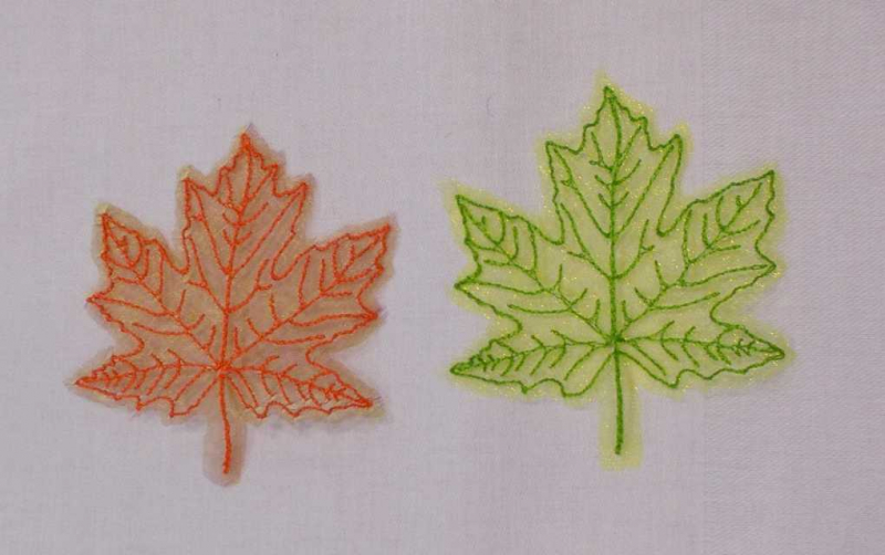 Stickdatei Set Herbstblätter inkl. ITH Anhänger, Foto mit 2 Blättern auf hellem Untergrund. Gestielte Blätter aus Organza, in orange oder grün, aufgestickte Blattaderstruktur selben Farbtons.