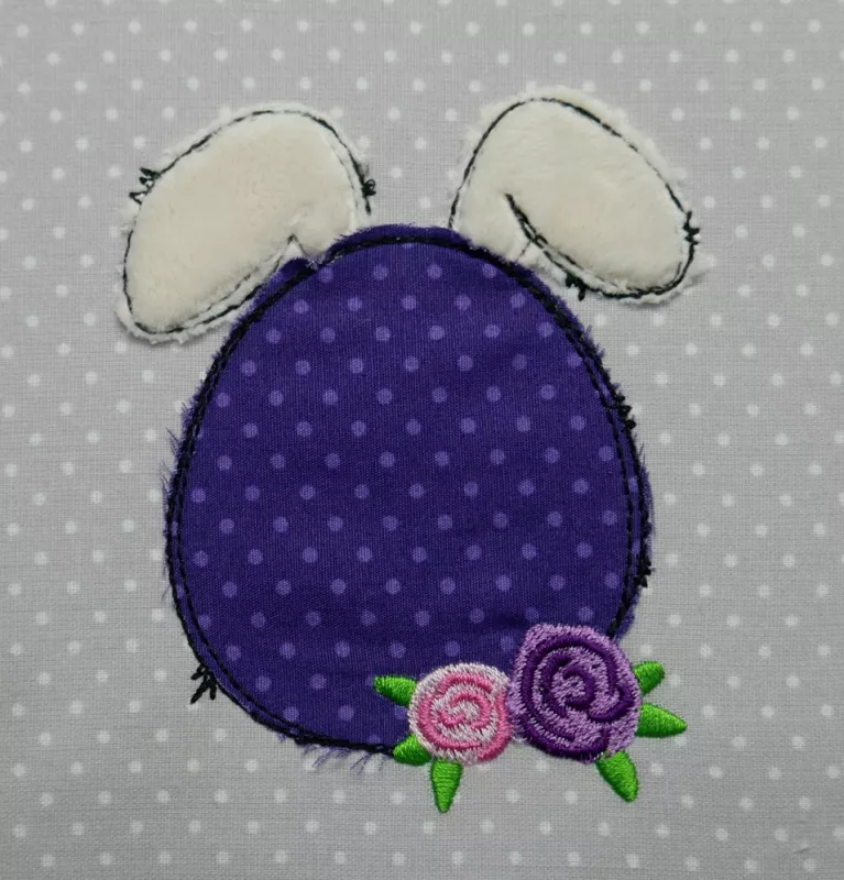 Stickdatei Set Oster-Doodles als Applikationen & Vollstick inkl. Schriftzüge, stilisierte Abbildung. Eiförmiger Kopf in lila mit heller lila  Punkten und hellen Flauschohren. Rosenblüten in rosa und pink am unteren Kopfrand.