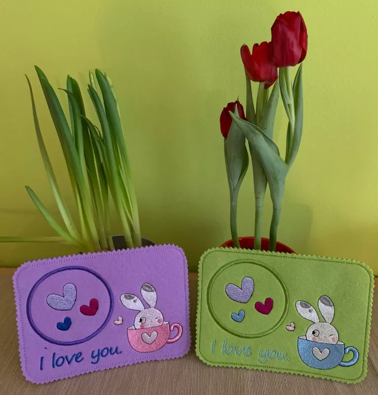 Freebie Stickdatei ITH Mug Rug Bunny in a Cup (10x10 & 13x18). Foto mit 2 Exemplaren im Querformat - eins auf lila und eins auf grünem Filzuntergrund - mit Motiv und Schriftzug in divers bunt, platziert vor zwei Topfblumen. Je kreisrunder Abstellplatz mit