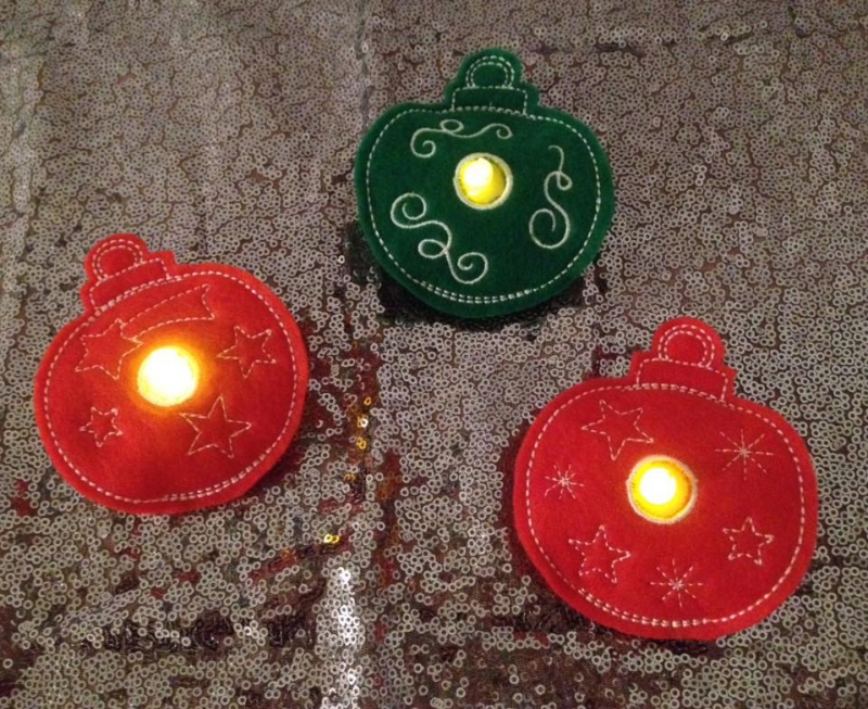 Stickdatei Set Christmas Lights (div. Rahmengrößen). Foto zeigt 3 LED-Cover in Form von Weihnachtsbaumkugeln, in jeder davon steckt mittig ein leuchtendes LED. Aus grünem, bzw. orangem Filz, filigran verziert.